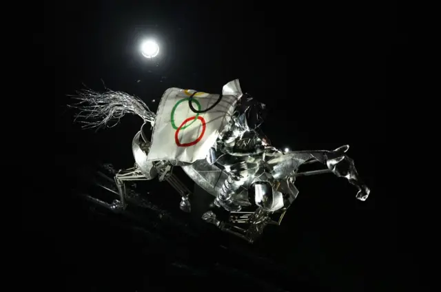 Una jinete vestida de plateado y con la bandera del Comité Olímpico Internacional en sus espaldas cabalga un caballo metálico sobre el río Sena durante la ceremonia inaugural de los Juegos Olímpicos París 2024.